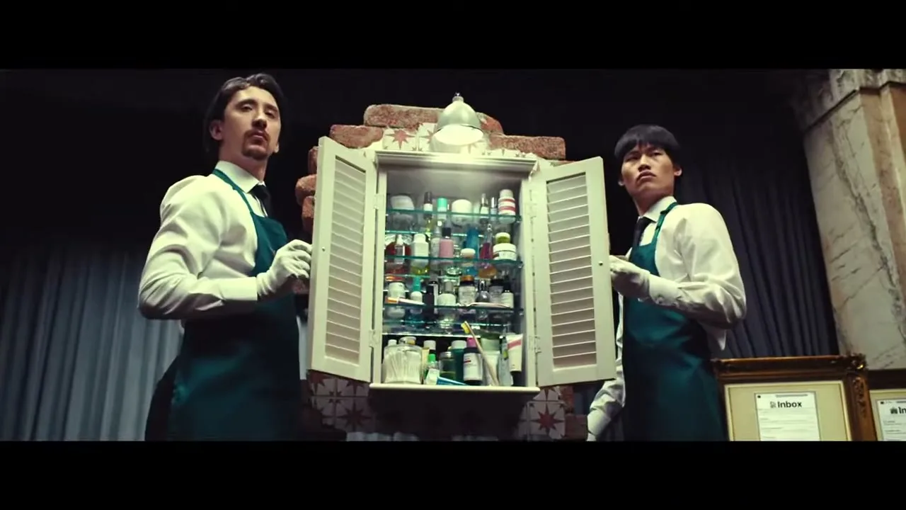 Two male attendants in white shirts and green aprons open a small cabinet revealing many personal items inside, in a dark, ornate room.