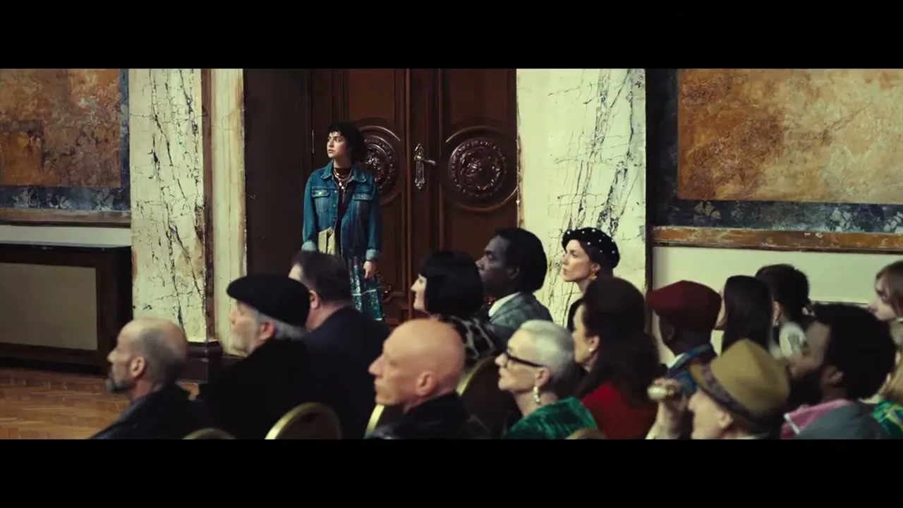 A young woman in a denim jacket stands in a doorway, looking up and to the left, while a diverse audience sits in rows facing away from her in an ornate hall.