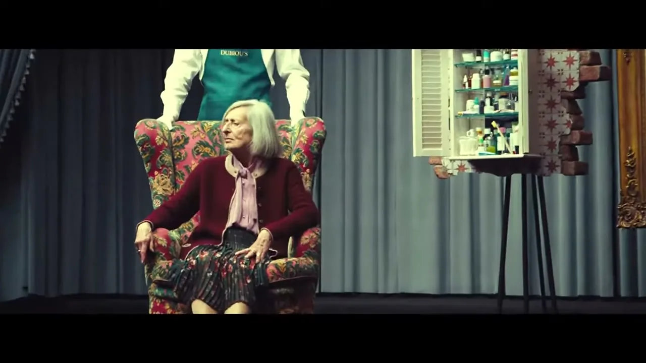 An elderly woman with white hair sits in a floral armchair on a stage, looking around as a man in a green apron stands behind her.