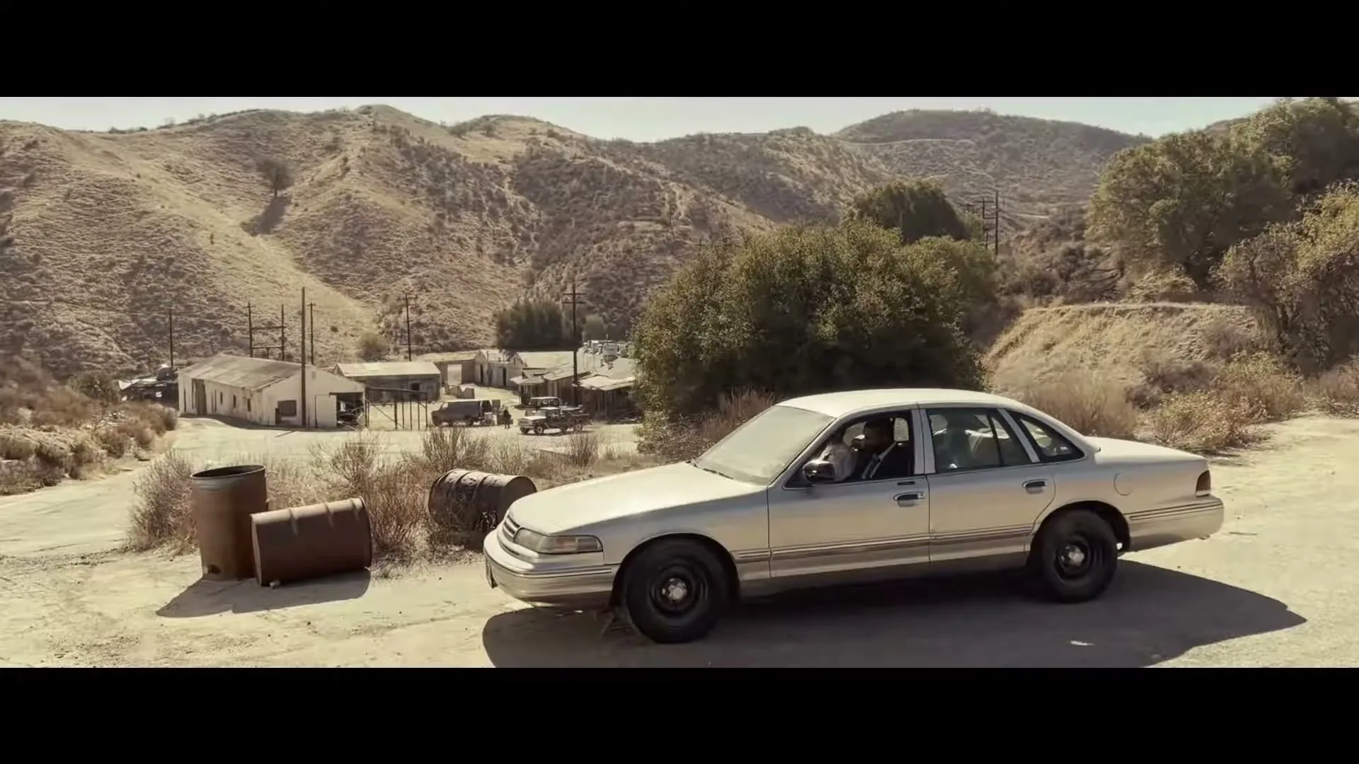 Two detectives in a silver sedan are parked on a dirt road overlooking a dusty, remote town in a valley surrounded by hills.