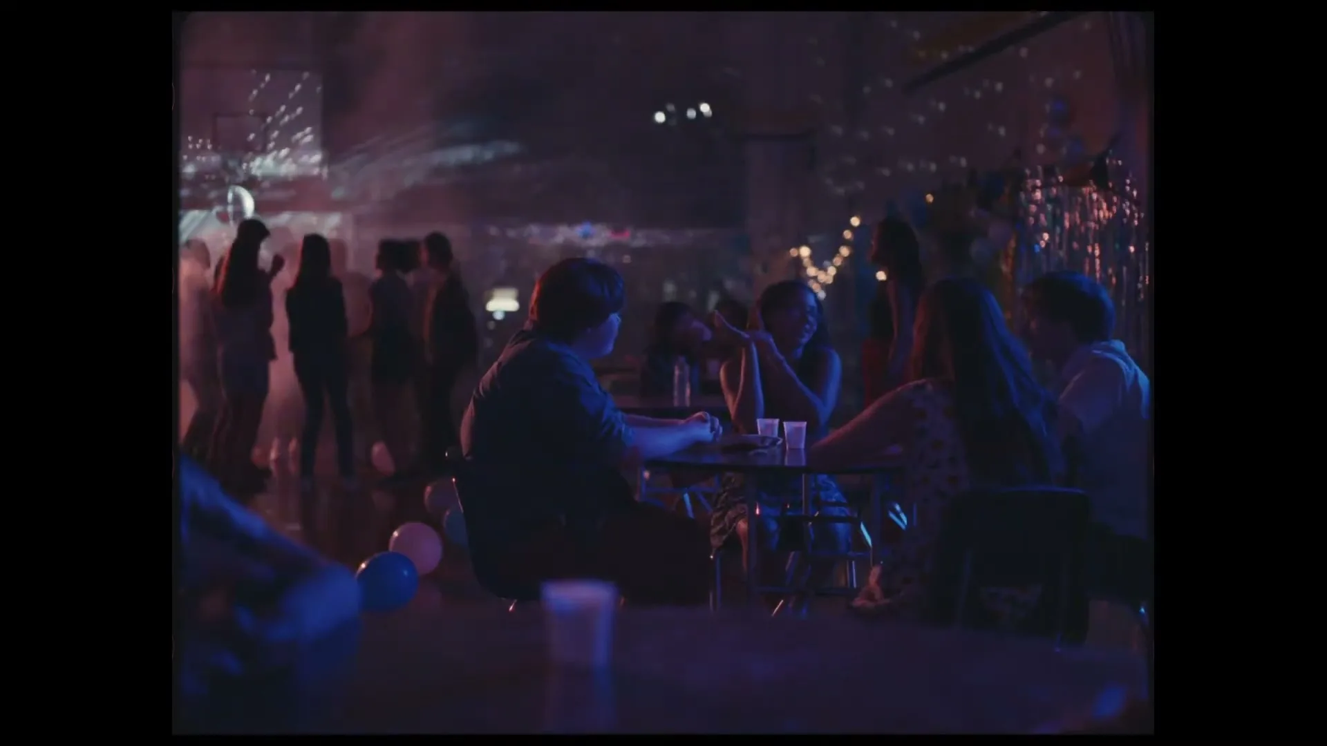 A young man sits alone at a table in a dimly lit, crowded party, looking around at other partygoers dancing and socializing.