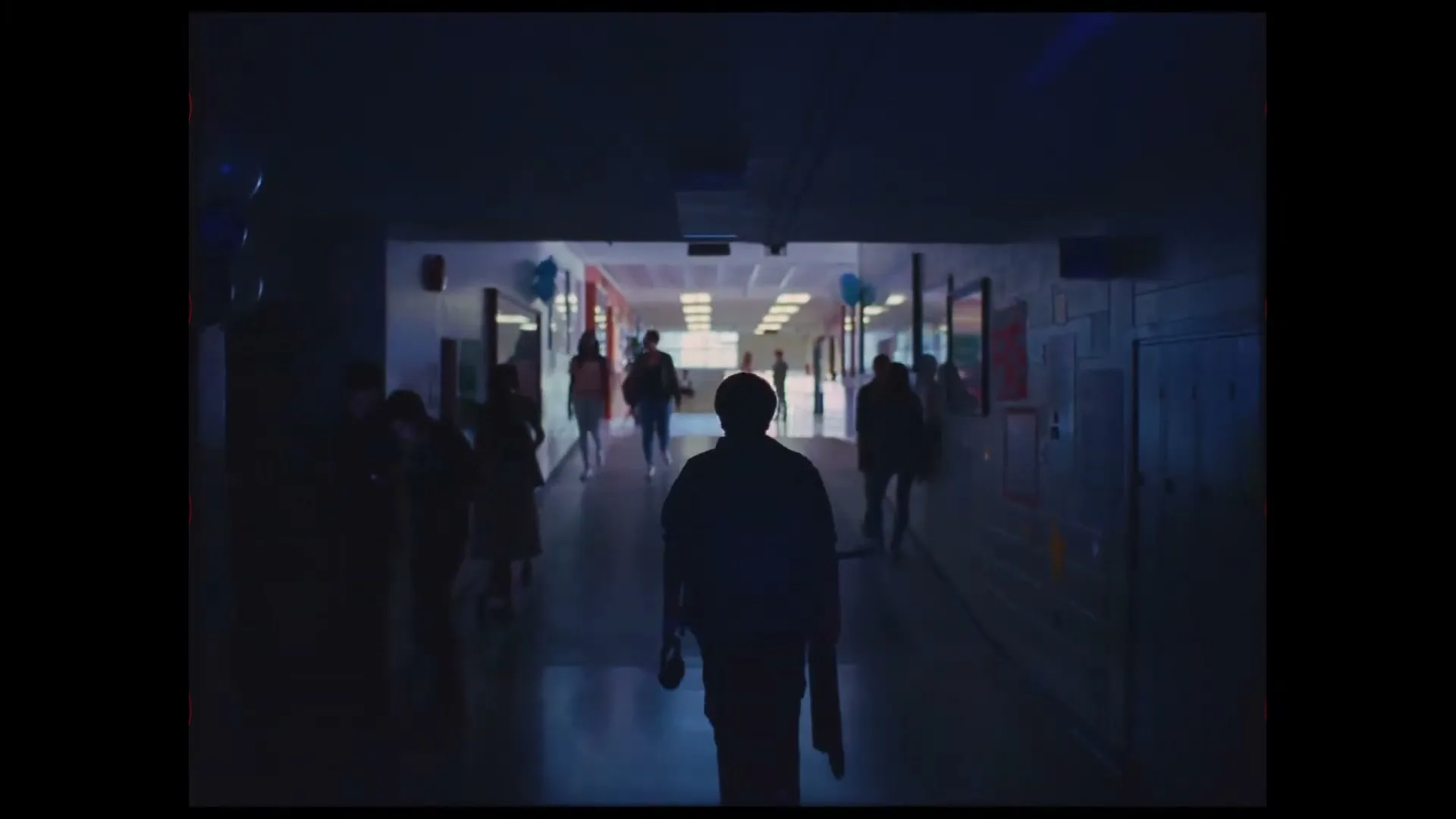 A young person walks down a dark school hallway, silhouetted against the brighter end, as other students walk past them.