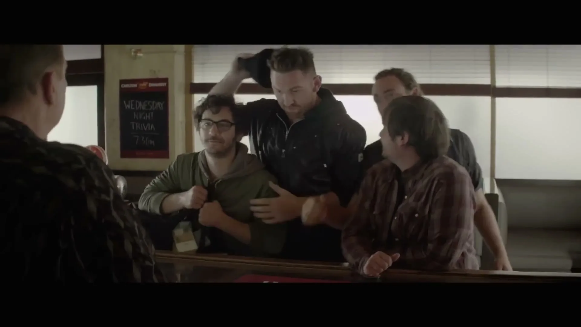 Four men stand at a bar, looking at a fifth man with varied expressions, in a dimly lit pub.