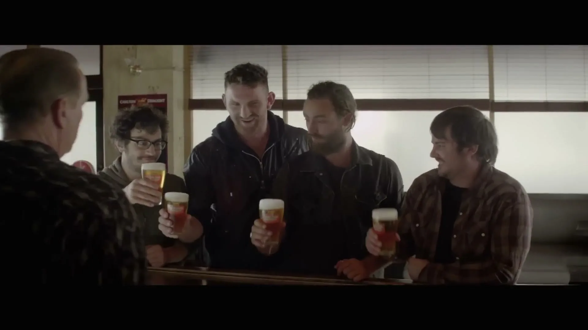 Four men at a bar raise their glasses of beer in a toast, smiling and looking at each other.