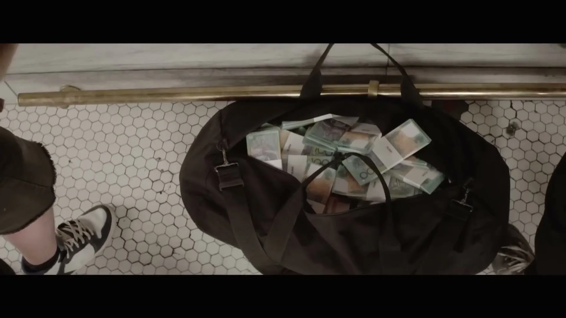 A black duffel bag overflowing with Australian banknotes sits on a white hexagonal tiled floor, next to a person's feet.