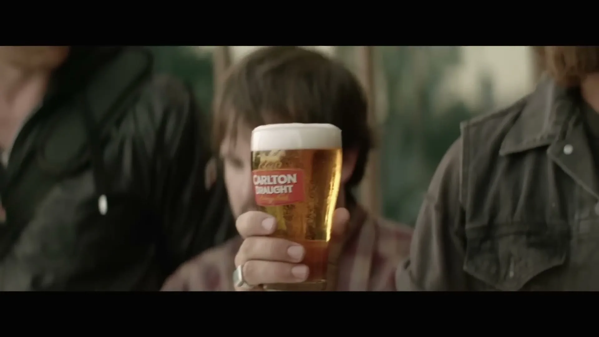 A man holds up a glass of Carlton Draught beer, with other men partially visible on either side, in a dimly lit interior setting.