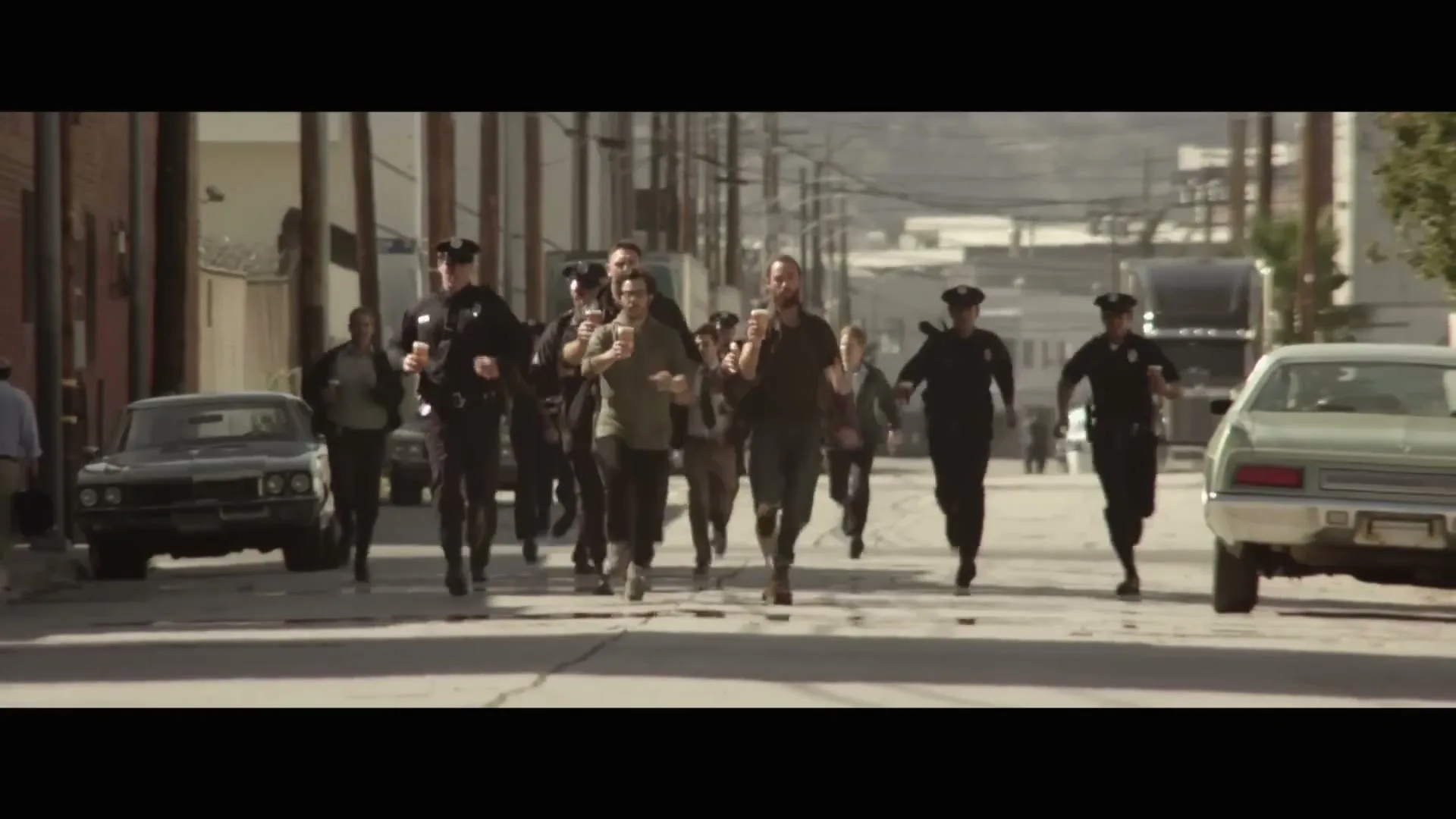 A large group of men, some dressed as police officers, run down a dusty street in an urban area, some holding cups.