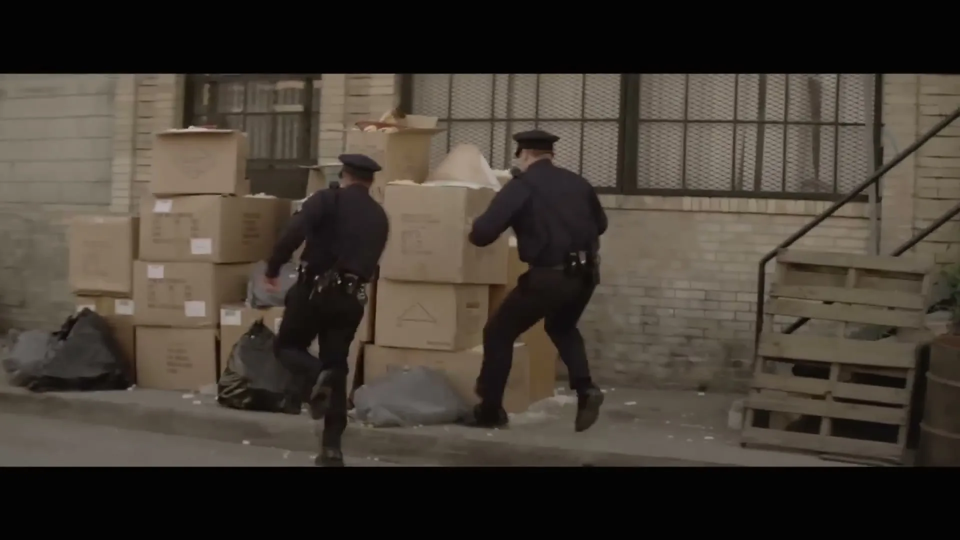 Two male police officers in uniform run through an alley, one of them crashes into a stack of cardboard boxes, sending white powder flying.