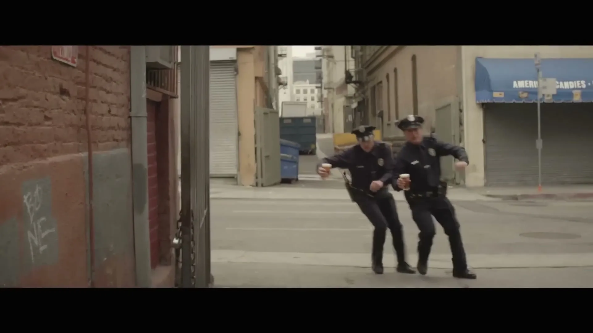 Two male police officers in uniform run awkwardly down a city street, holding coffee cups, with brick walls and buildings in the background.