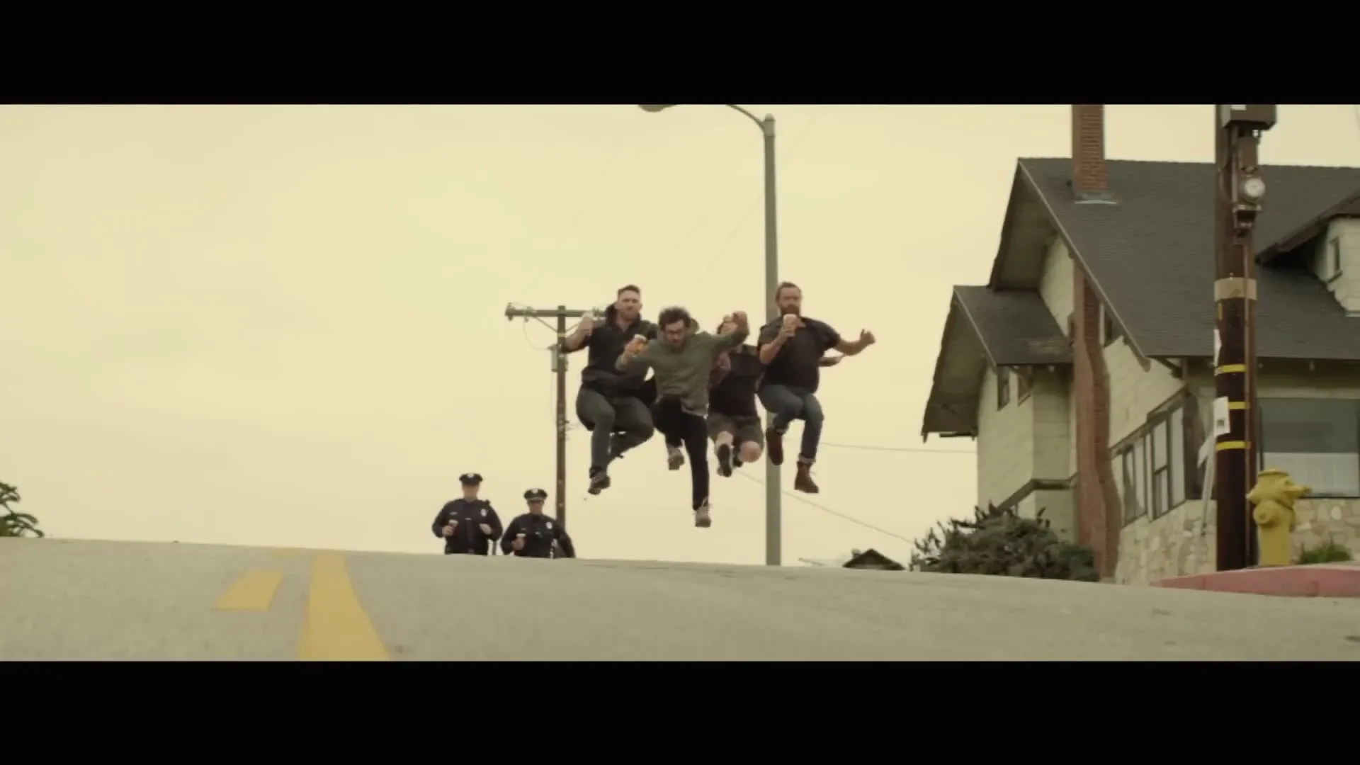 Five men in casual clothes run up a hill, holding beers, with two police officers chasing them in the background on a cloudy day.
