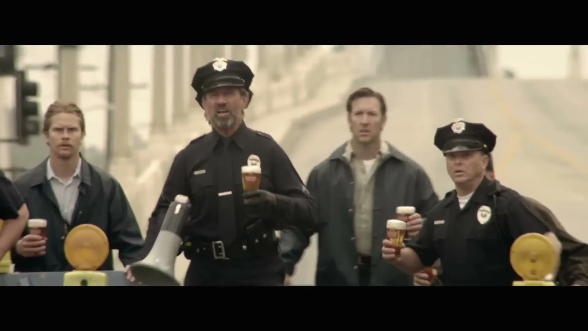 A group of men, some dressed as police officers, hold beers and a megaphone while walking on a bridge during the day.