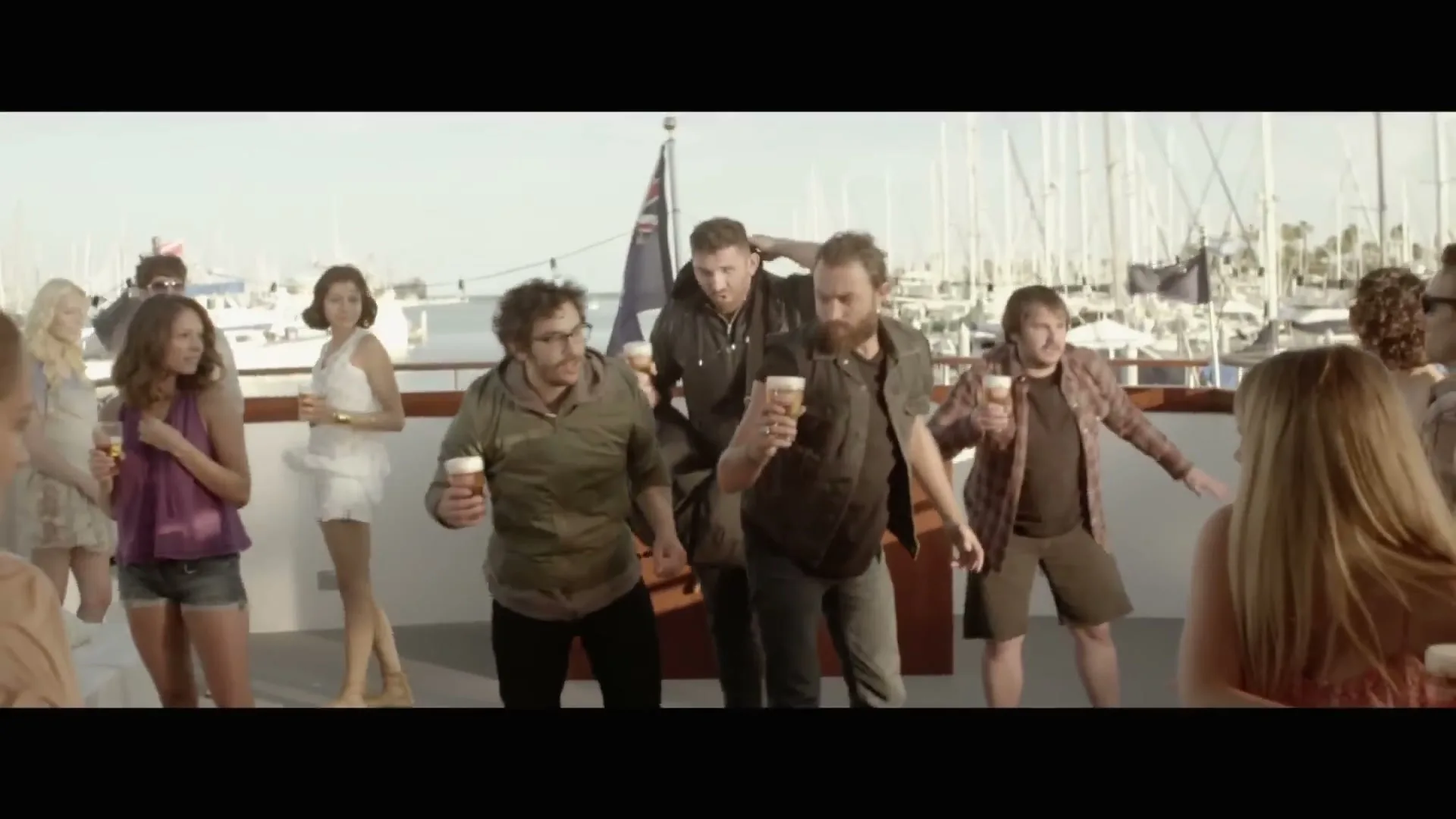 A group of men and women are on a boat, holding beers, as the men move in a synchronized, slightly awkward dance, with sailboats in the background.