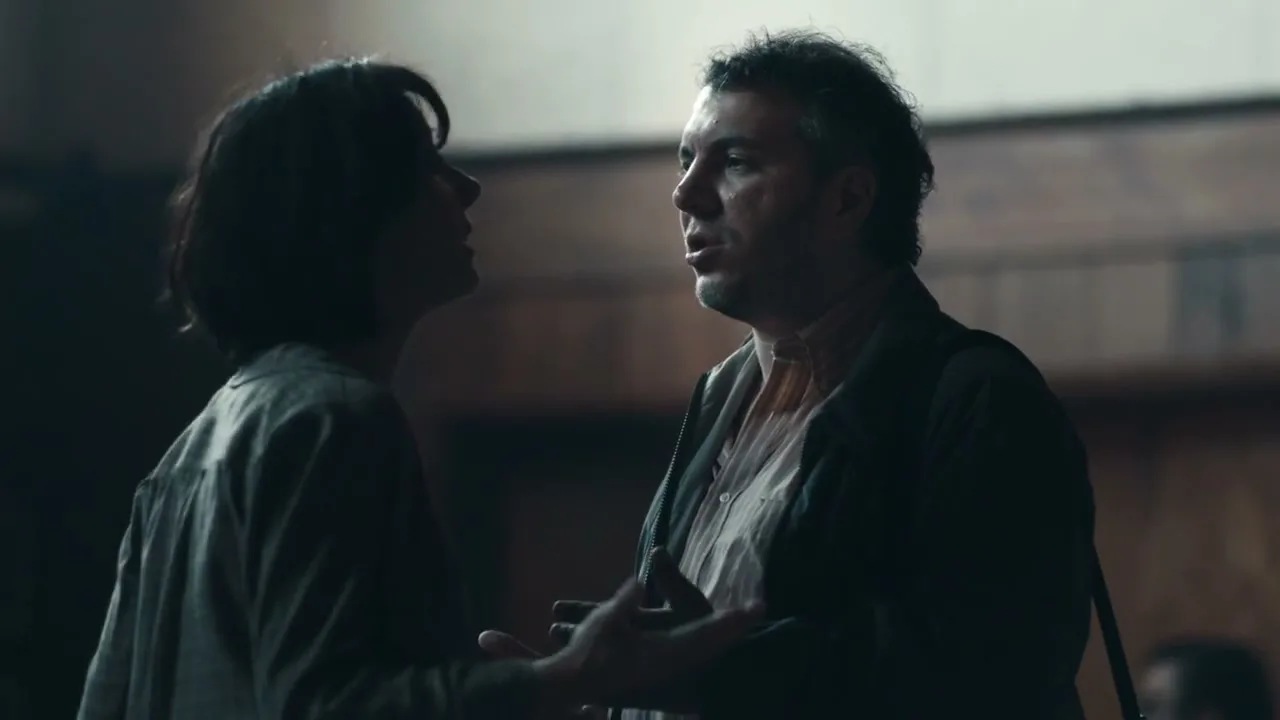 A man and a woman, both in their 30s, are having an intense conversation in a dimly lit interior space.