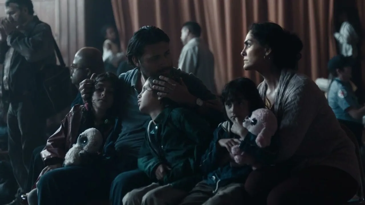 A family of five, including two parents and three children, sits together in a crowded shelter, looking up with worried expressions.