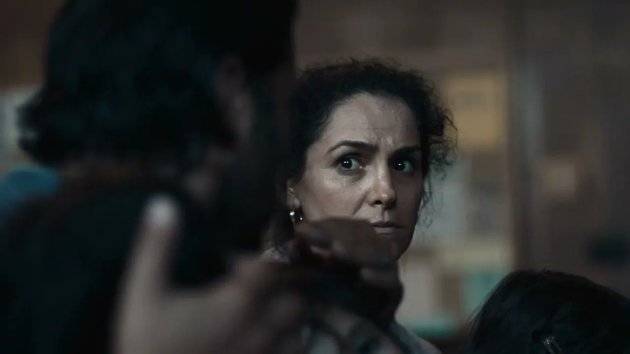 A distressed woman with dark curly hair looks over a man's shoulder, her eyes wide with worry in a dimly lit interior.