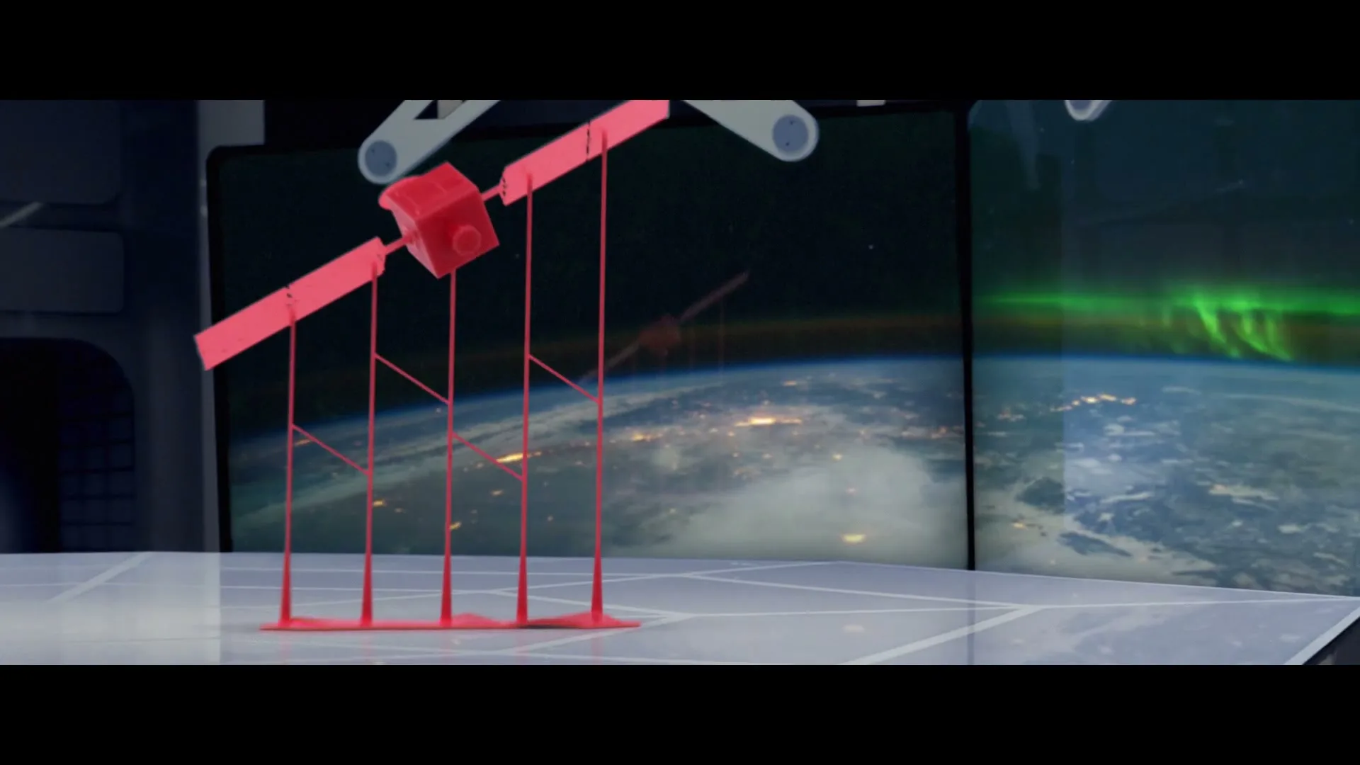 A red 3D-printed model of a satellite is on a white surface, with two screens behind it showing Earth from space at night.