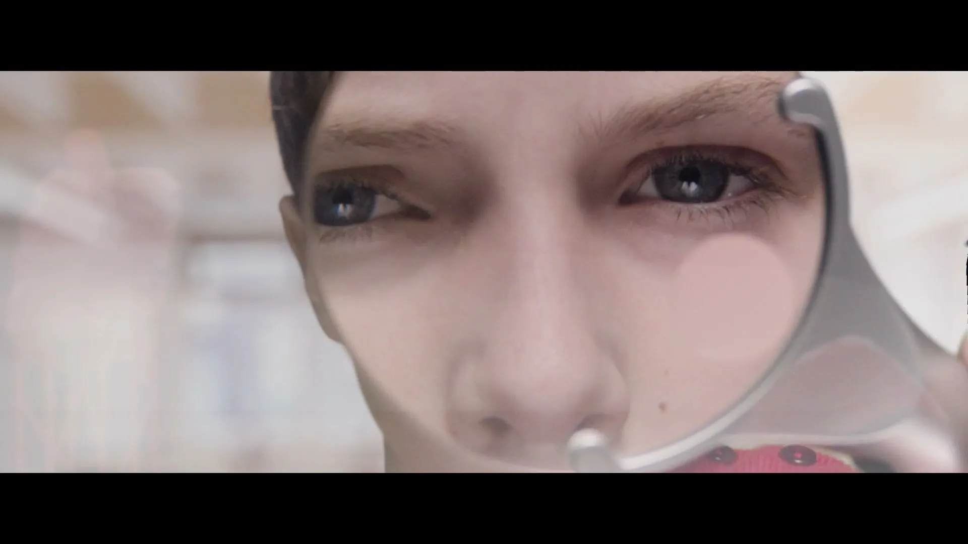 A young person with light skin and blue eyes looks directly at the camera, holding a magnifying glass to their face, in an interior setting.