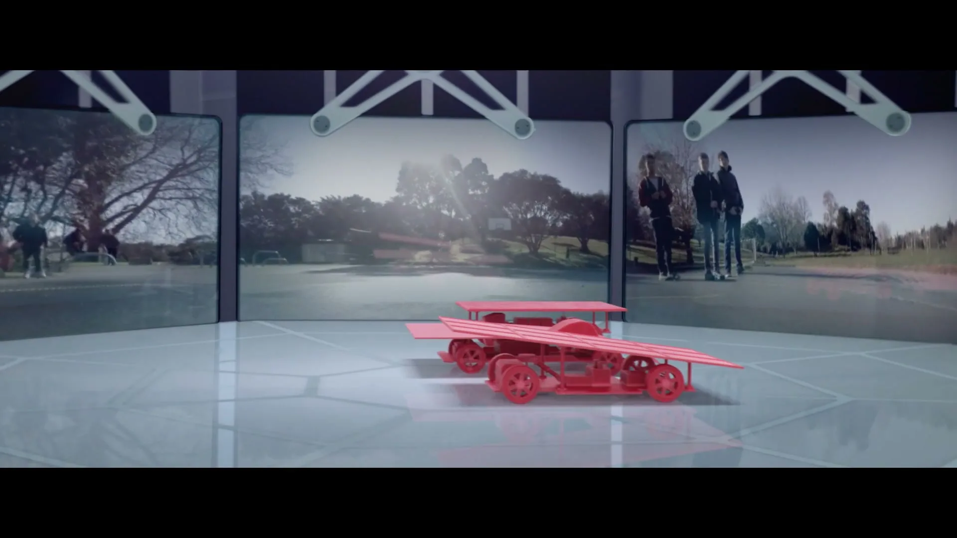 A red 3D-printed car is assembled on a glossy white surface, with three screens in the background showing outdoor scenes with people.