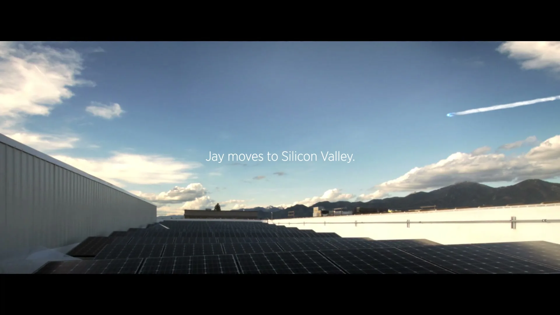 A wide shot of solar panels on a rooftop under a blue sky with mountains in the distance, as a plane flies overhead.