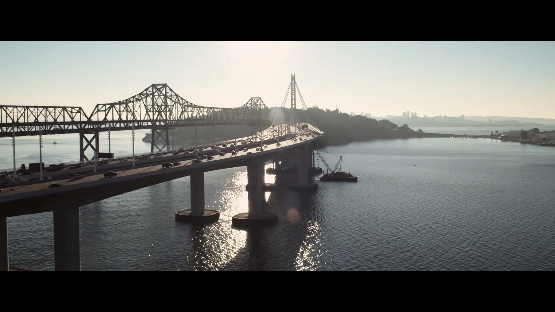 An aerial view captures a long bridge with cars driving across it, spanning a wide body of water under a bright sky.