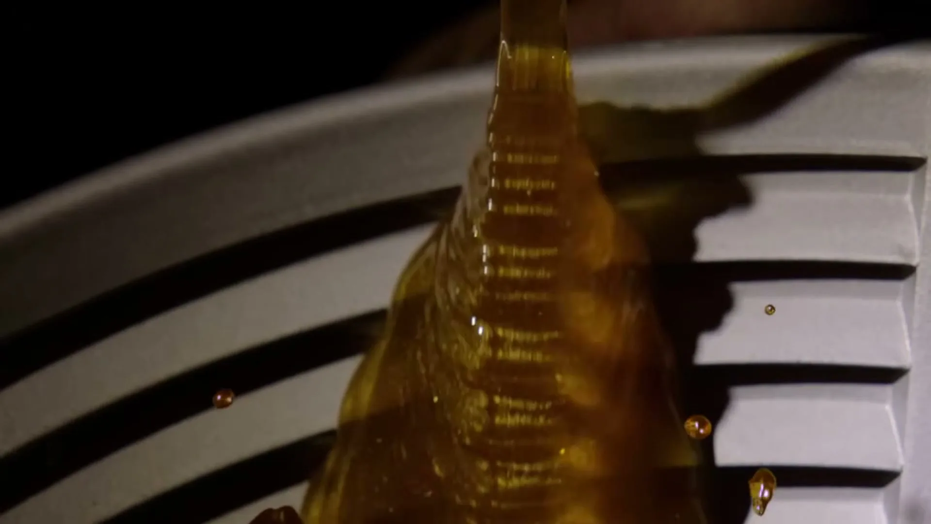 A thick, golden liquid pours onto a white, ridged surface, creating ripples and small splashes in a dark setting.
