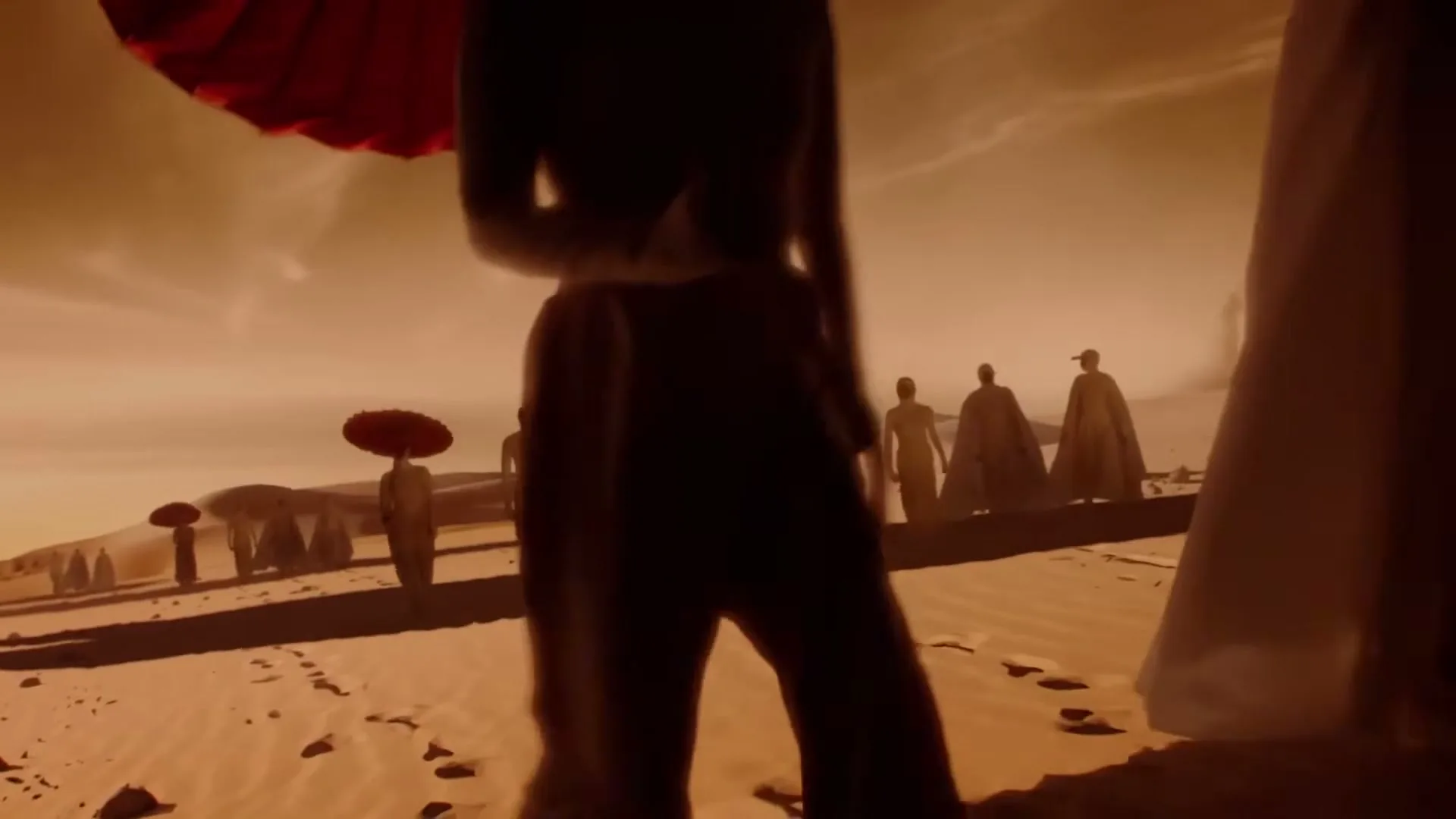 A group of people in flowing robes and carrying red parasols walk across a vast, sandy desert under an orange sky.