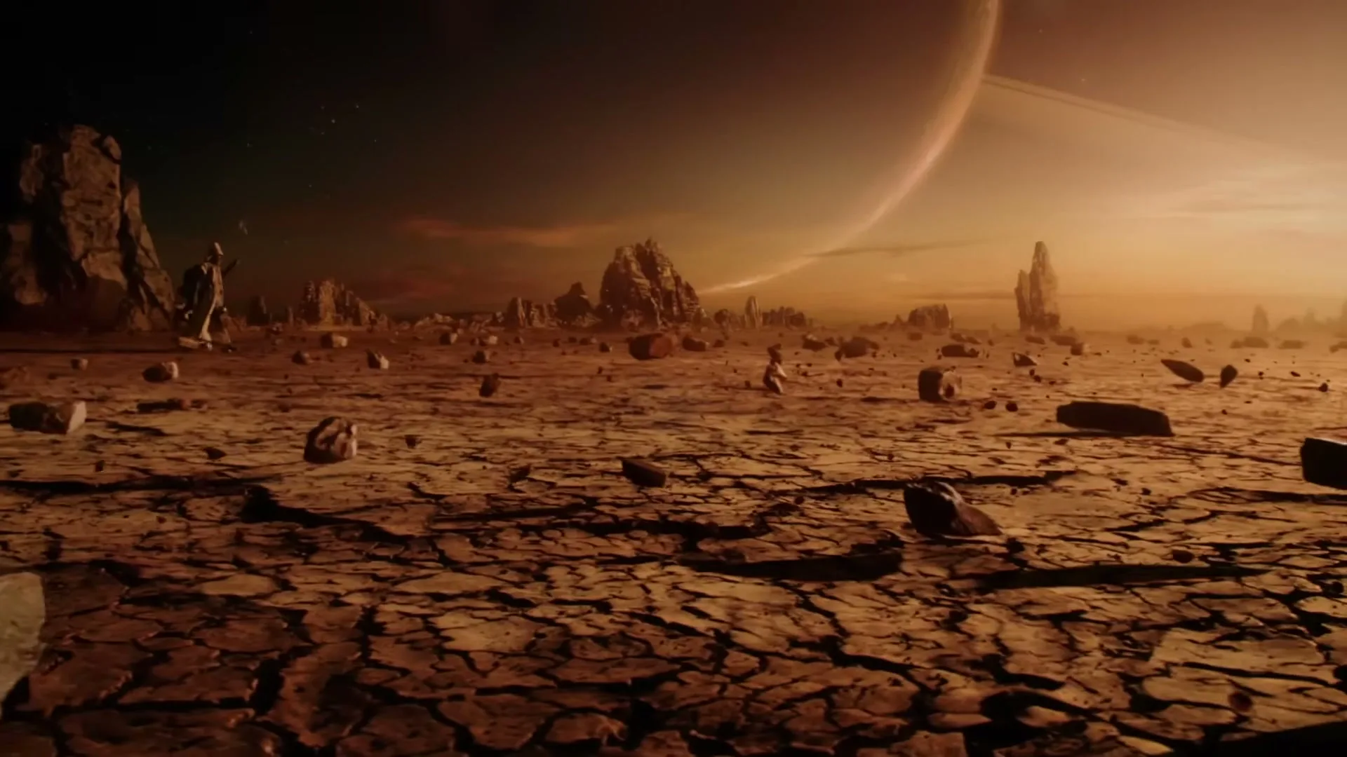 A wide shot of a desolate, cracked alien landscape with large rock formations and a ringed planet visible in the sky.
