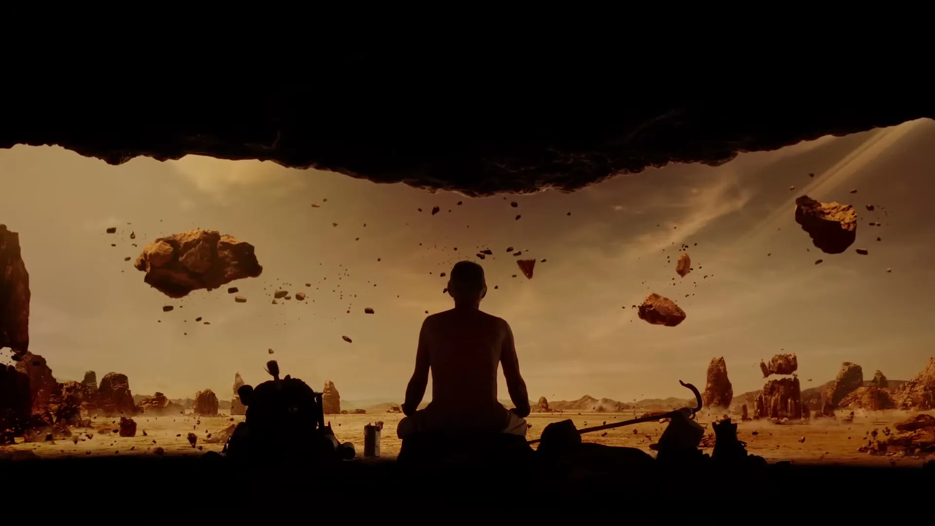 A man meditates in a desert landscape with floating rocks, seen from inside a cave.