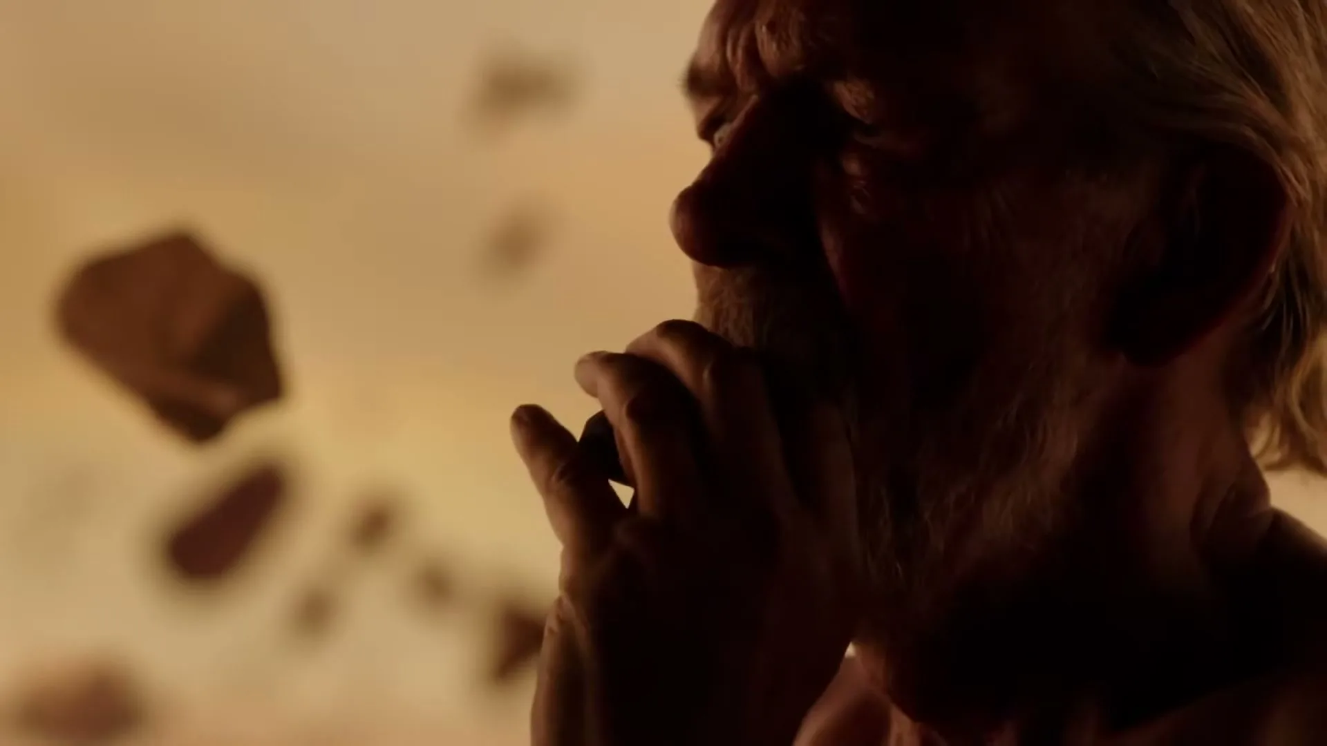 A silhouetted older man with a beard brings his hands to his mouth, looking pensive, with floating debris in a warm, ethereal space.