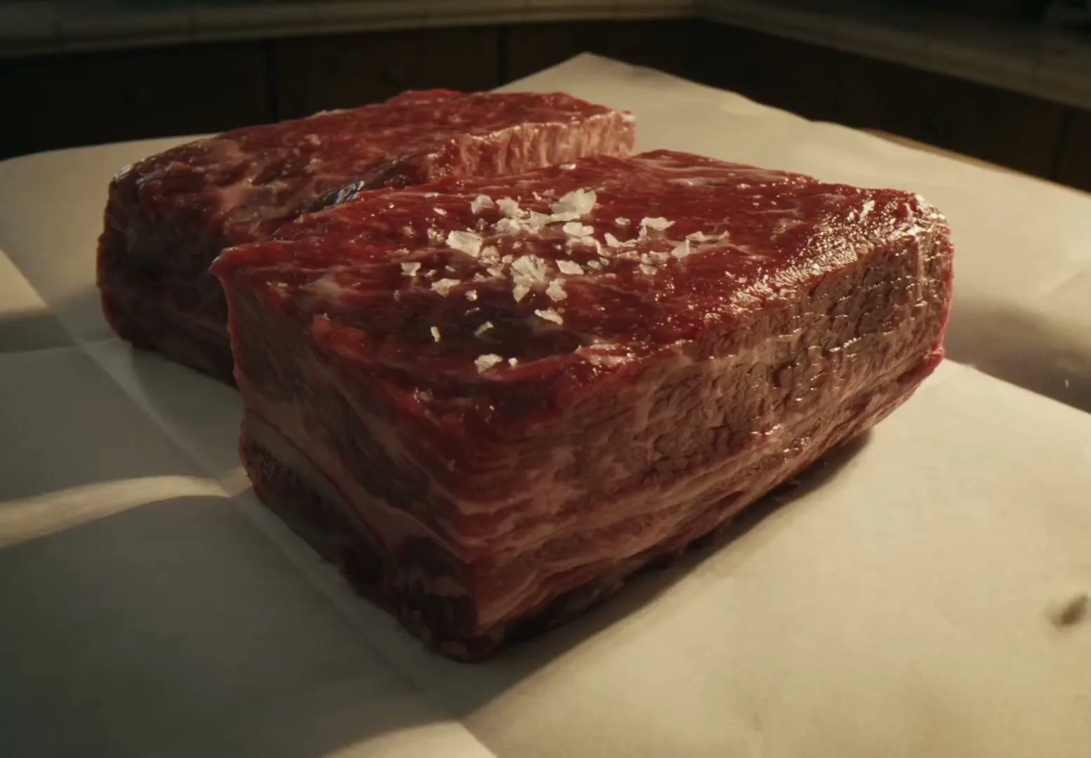 Two raw short ribs, seasoned with coarse salt, sit on white parchment paper on a kitchen counter, ready for cooking.
