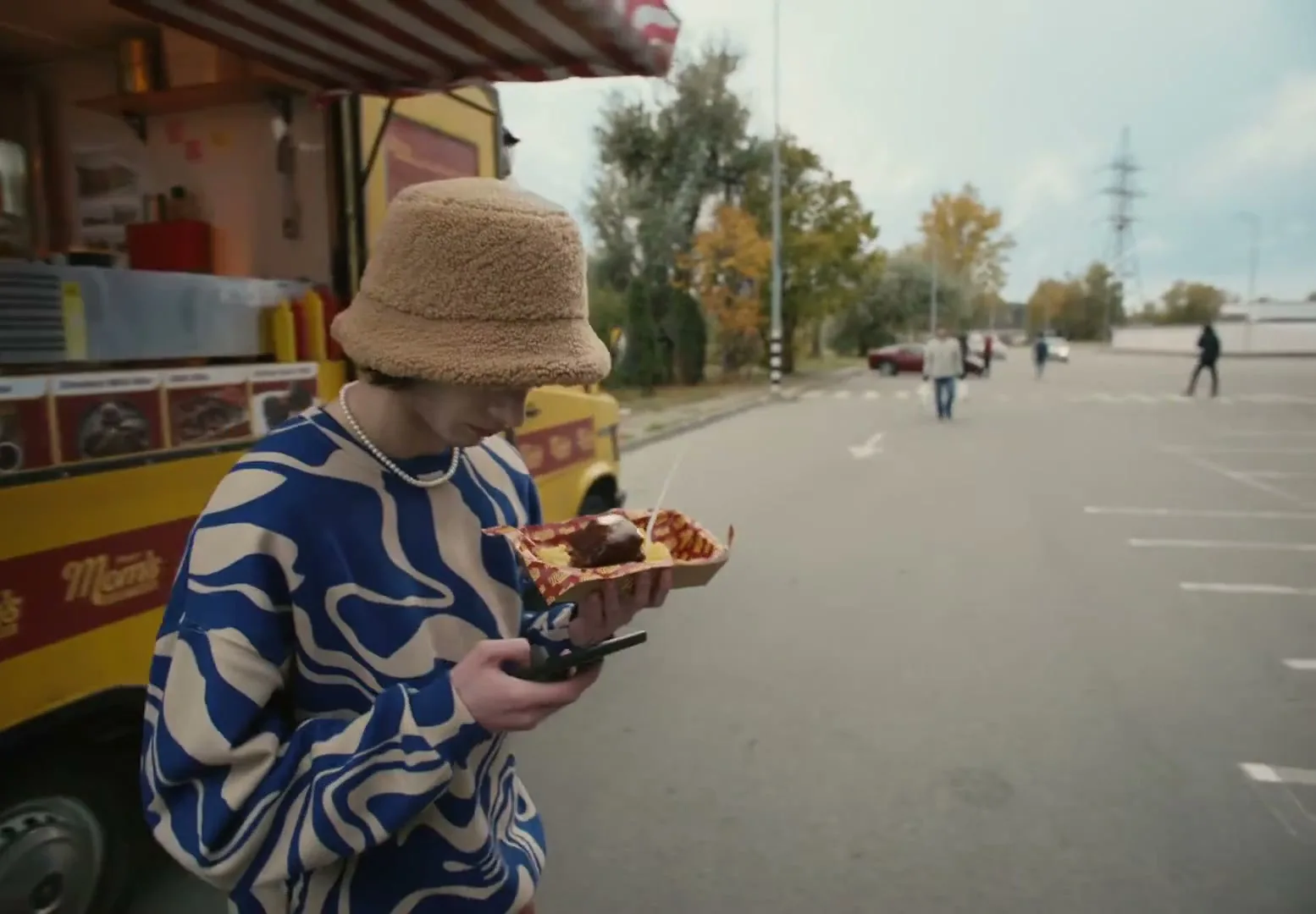 A young person in a bucket hat and patterned sweater receives food from a vendor at a food truck, then takes a selfie with the food in a parking lot.