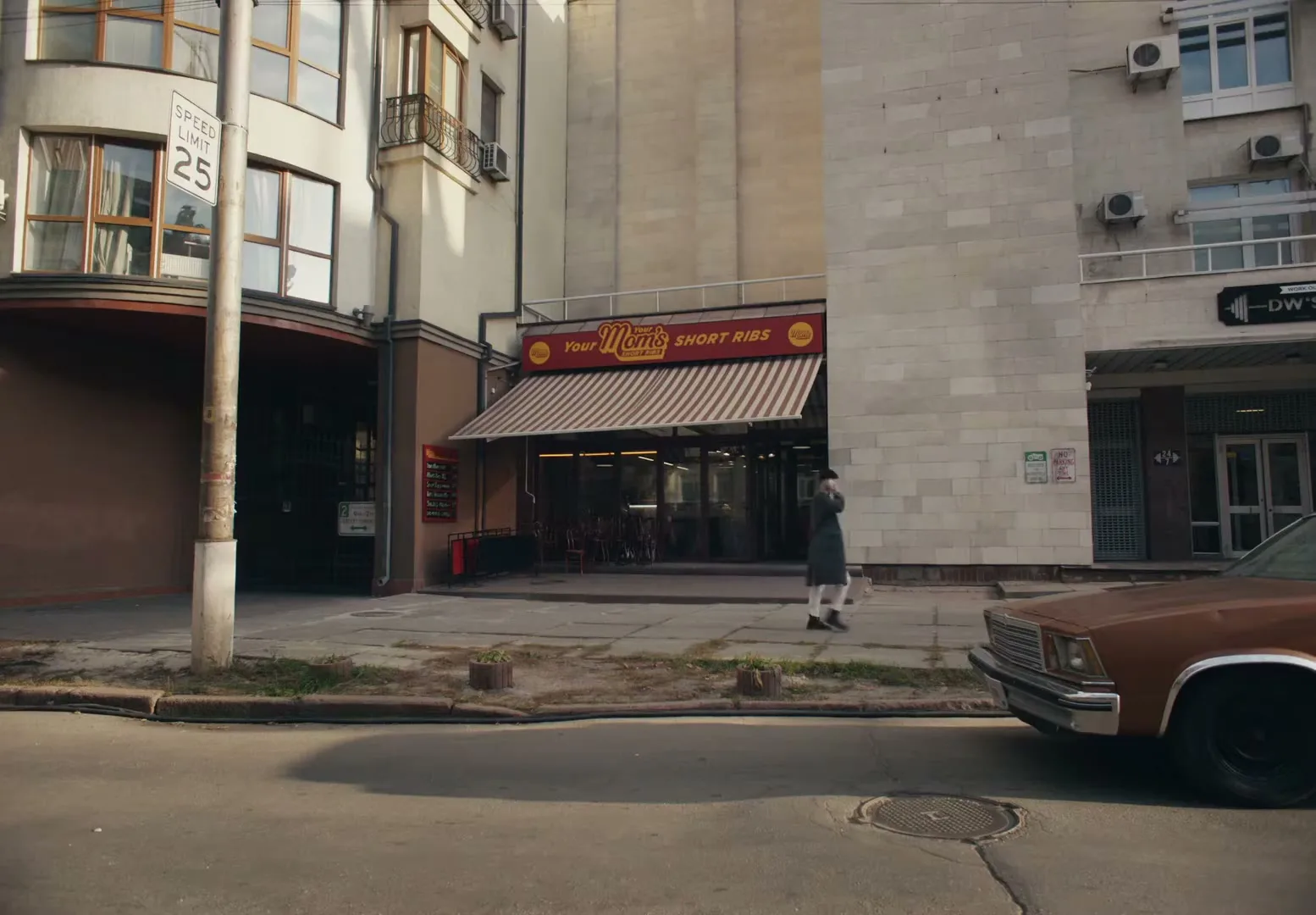 A person in a dark coat walks past a restaurant with a striped awning that reads "Your Mom's Short Ribs" on a city street.