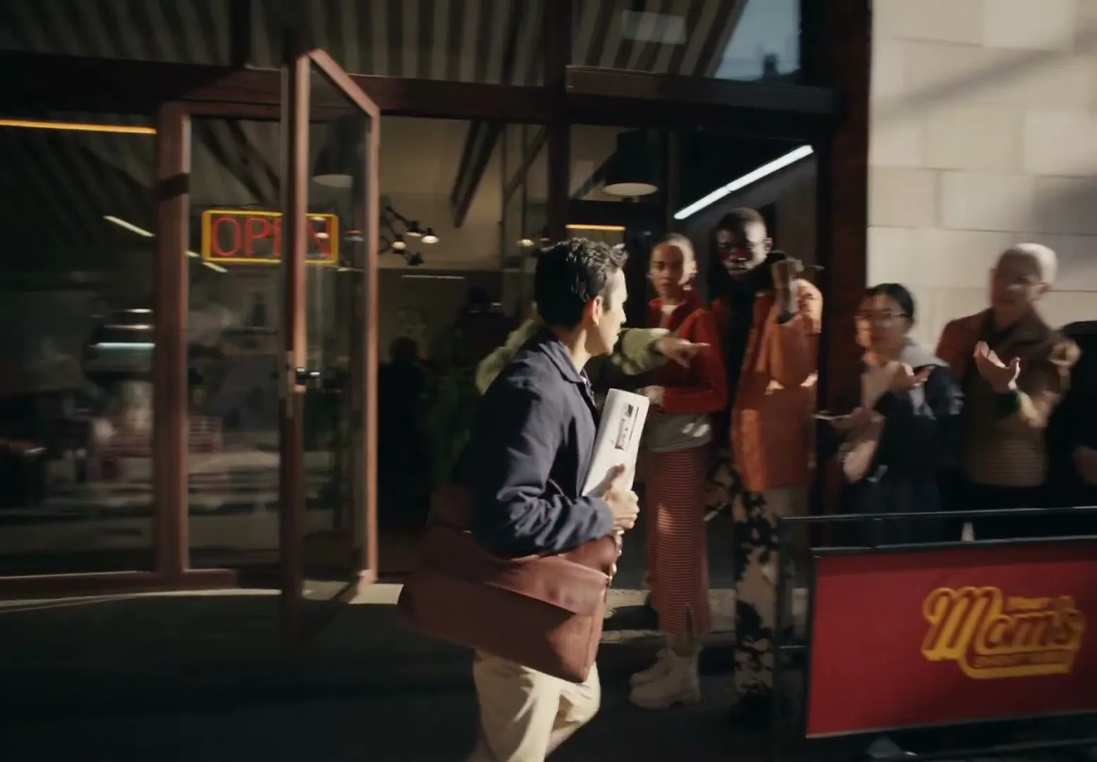A man in a blue jacket and khaki pants walks past a long line of diverse people waiting outside a restaurant called "Your Mom's Short Ribs" on a sunny day.