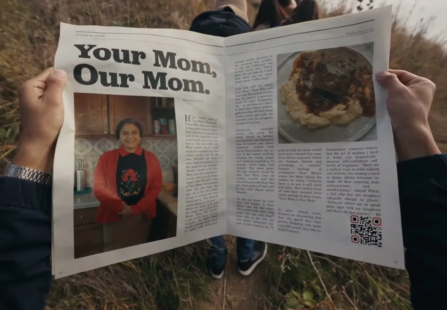 A person holds up a newspaper with the headline "Your Mom, Our Mom" and an image of a woman in a kitchen, while standing in a grassy field.