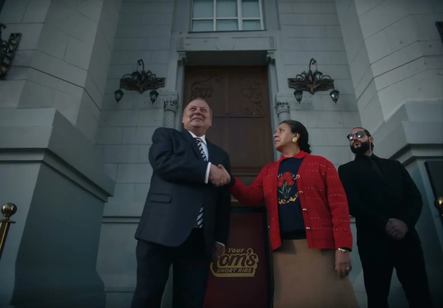 A man in a suit and a woman in a red cardigan shake hands in front of a building, with a bodyguard standing nearby.