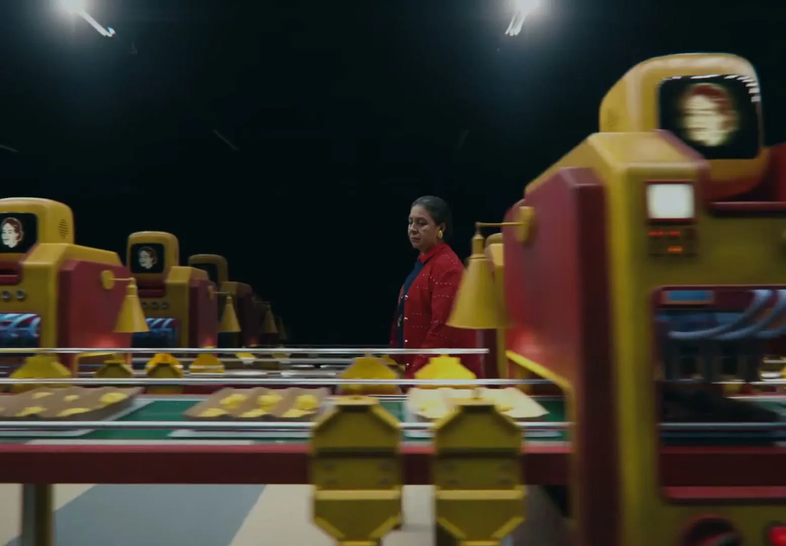 A woman in a red jacket walks through a factory of cartoonish robots with screens for heads, as food moves along a conveyor belt.
