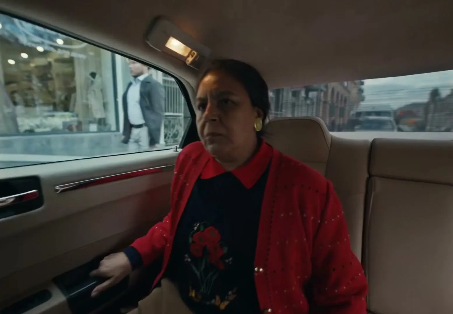 An older woman with dark hair and a red cardigan gets into the back seat of a car, looking around with a neutral expression.