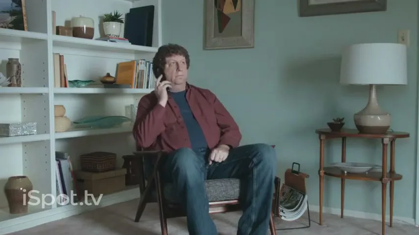 A man with curly brown hair, wearing a red shirt and jeans, sits in a chair in a living room, holding a phone to his ear.