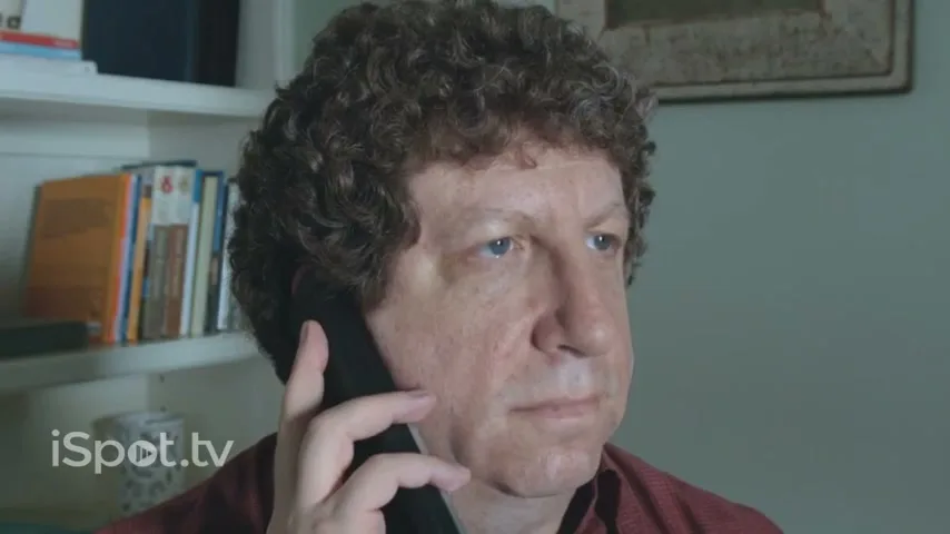 A middle-aged man with curly hair holds a phone to his ear, looking bored while on hold in a dimly lit room.