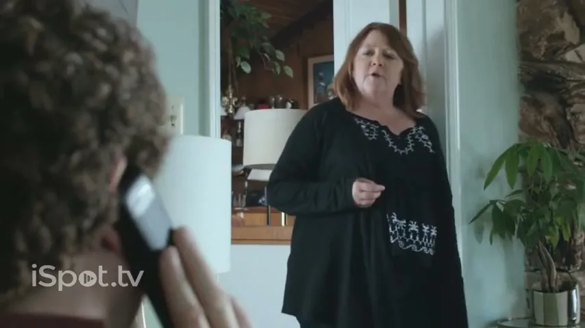 A woman in a black top stands in a doorway, looking at a man with curly hair who is on the phone, in a home interior.