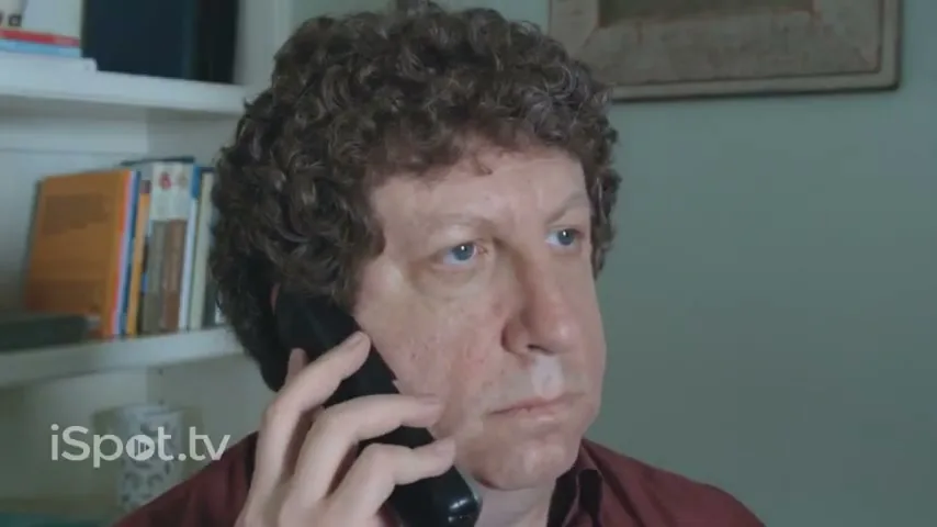 A middle-aged man with curly hair holds a phone to his ear, looking annoyed while on hold in a home office.
