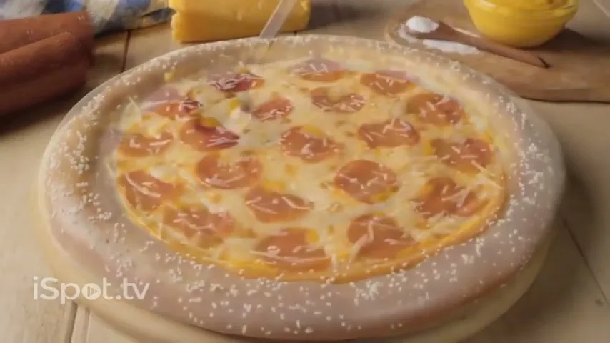 A pizza is being assembled on a wooden surface, with a ladle spreading orange cheese sauce on the dough, then pepperoni and mozzarella appear, and finally the crust is covered in sesame seeds.