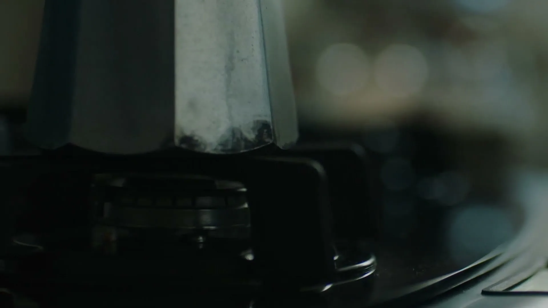 A close-up shot shows a silver moka pot on a black gas stove, as a blue flame ignites beneath it in a kitchen.