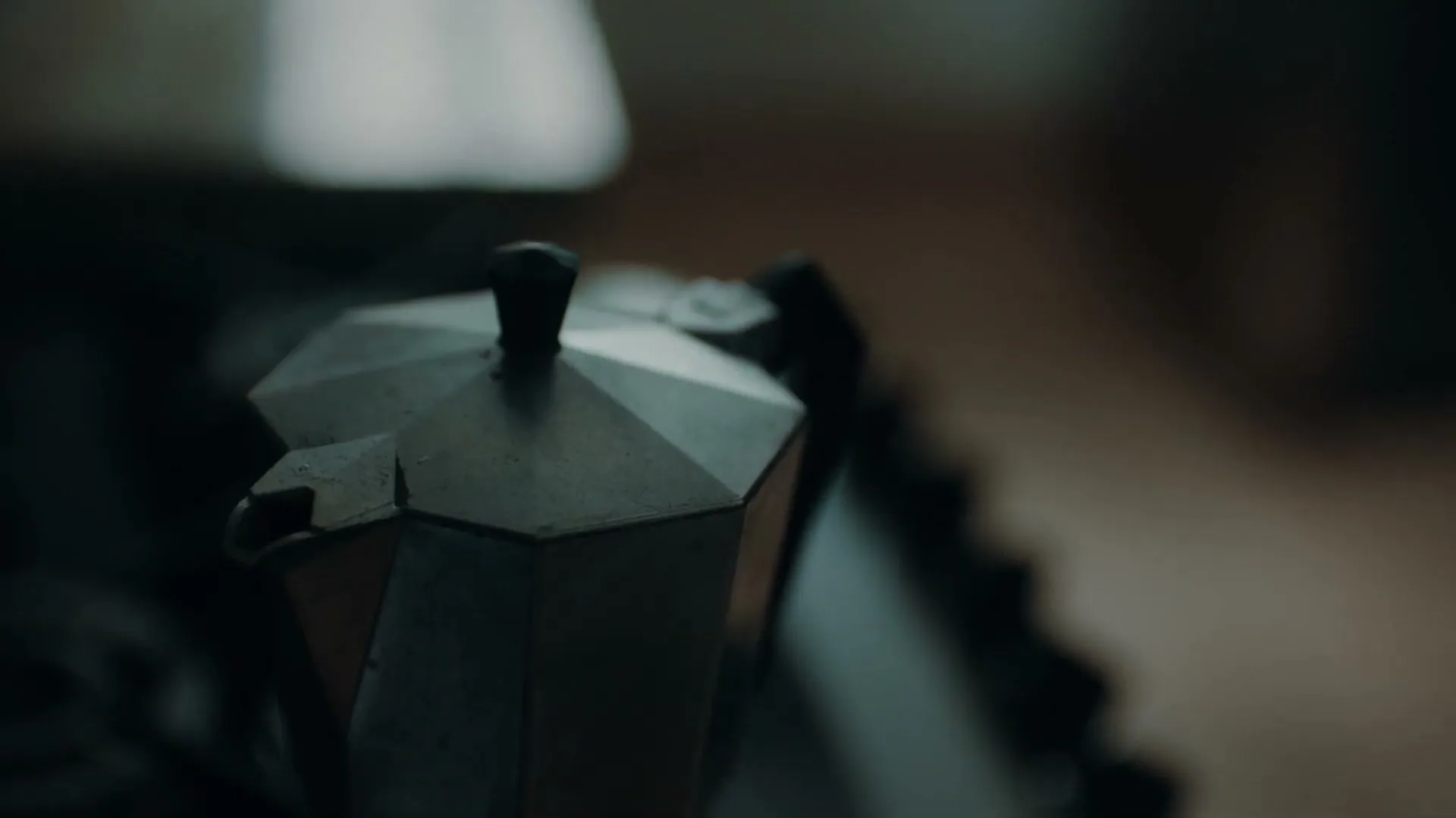 A close-up shot of a moka pot on a stove, with steam gently rising from its spout in a dimly lit kitchen.