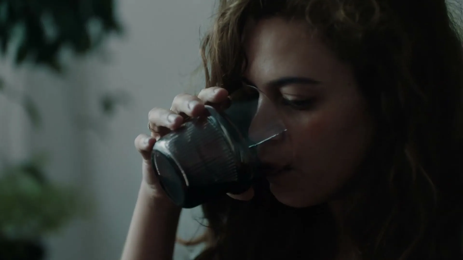 A young woman with curly brown hair and a nose ring drinks from a glass in a dimly lit room.