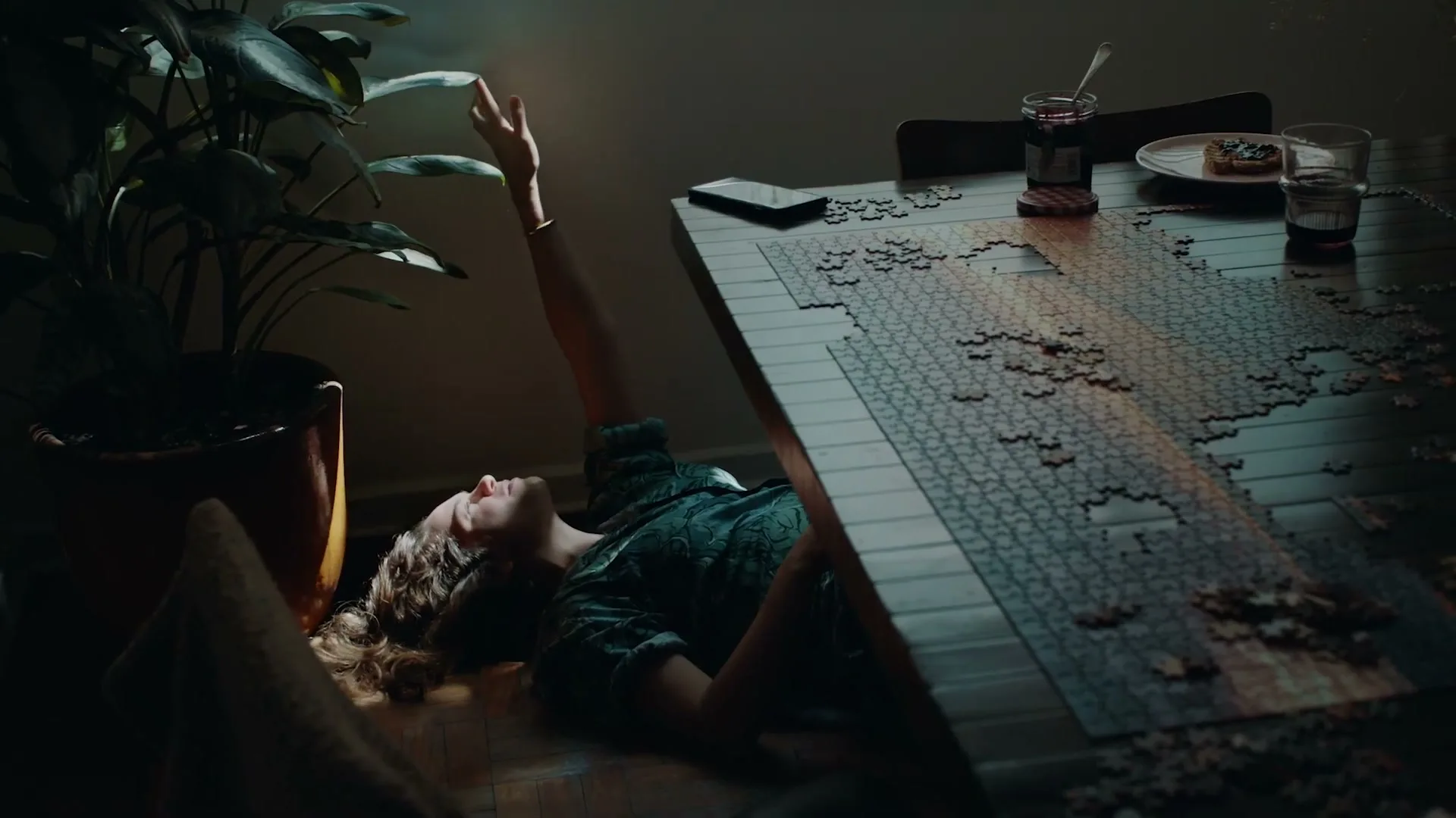 A young woman in pajamas lies on the floor, reaching up to a sunbeam, next to a table with a half-finished puzzle and breakfast items.