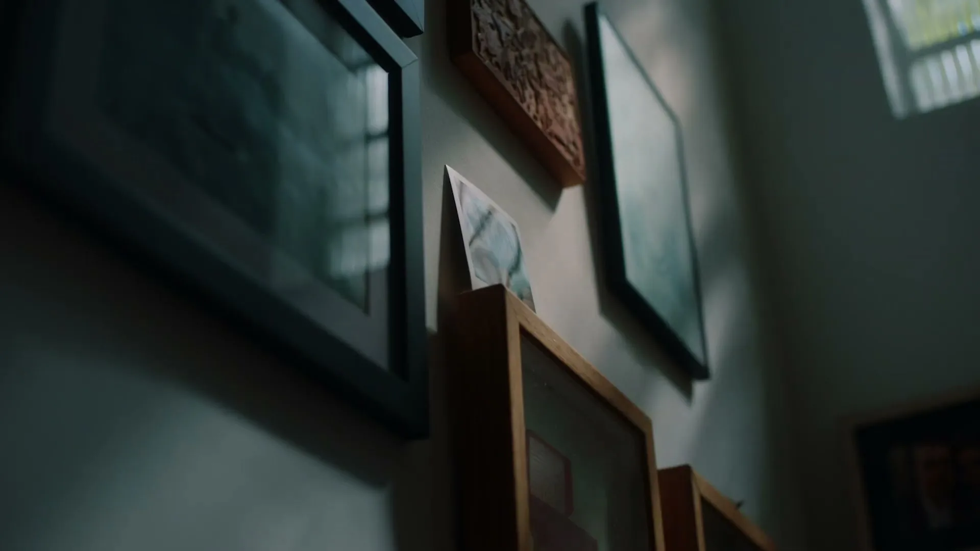 A low angle shot shows framed pictures and artwork hanging on a wall, with sunlight streaming through a window on the right.