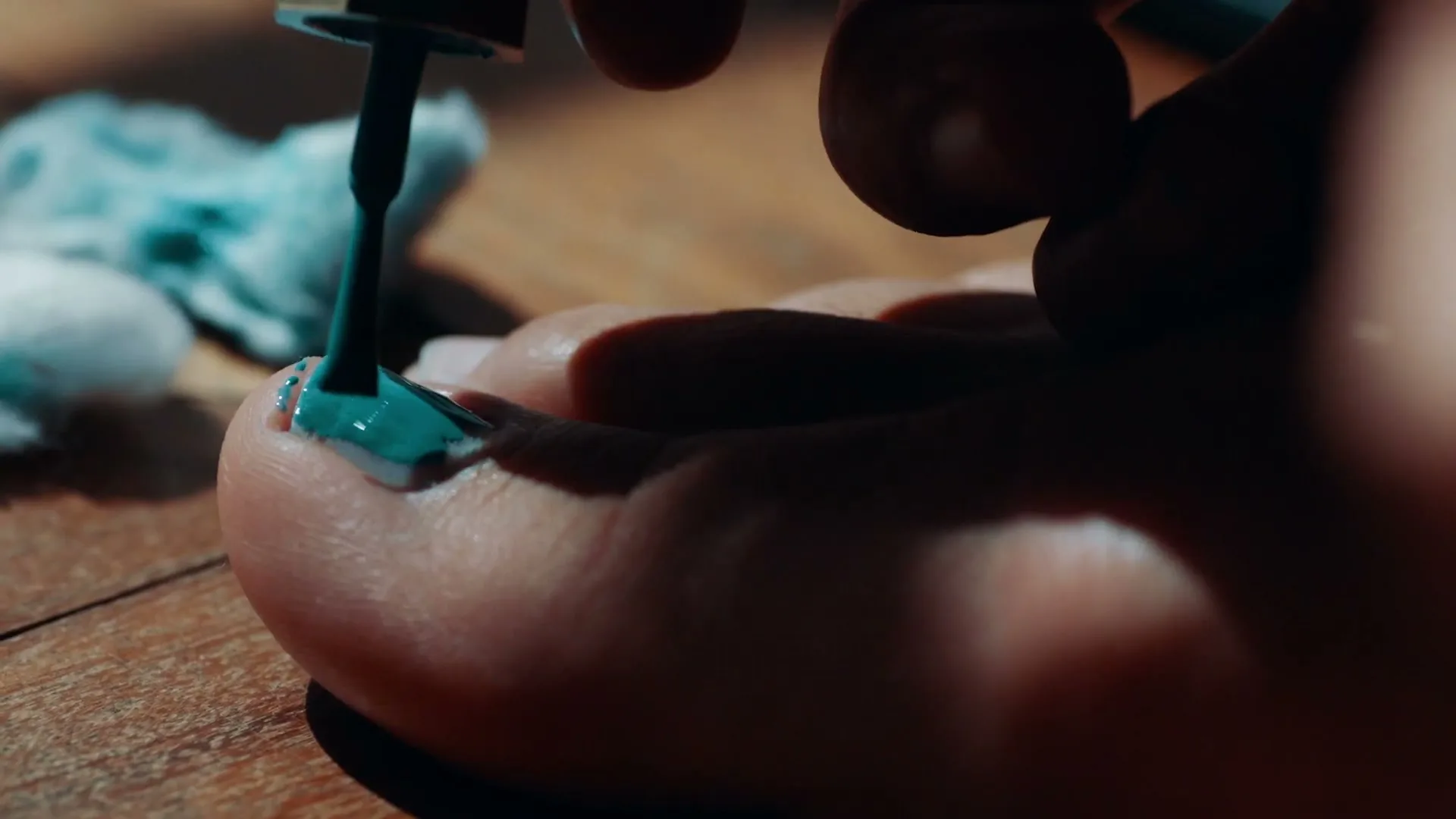 A person's foot is shown as they paint their toenails with bright blue nail polish on a wooden floor.