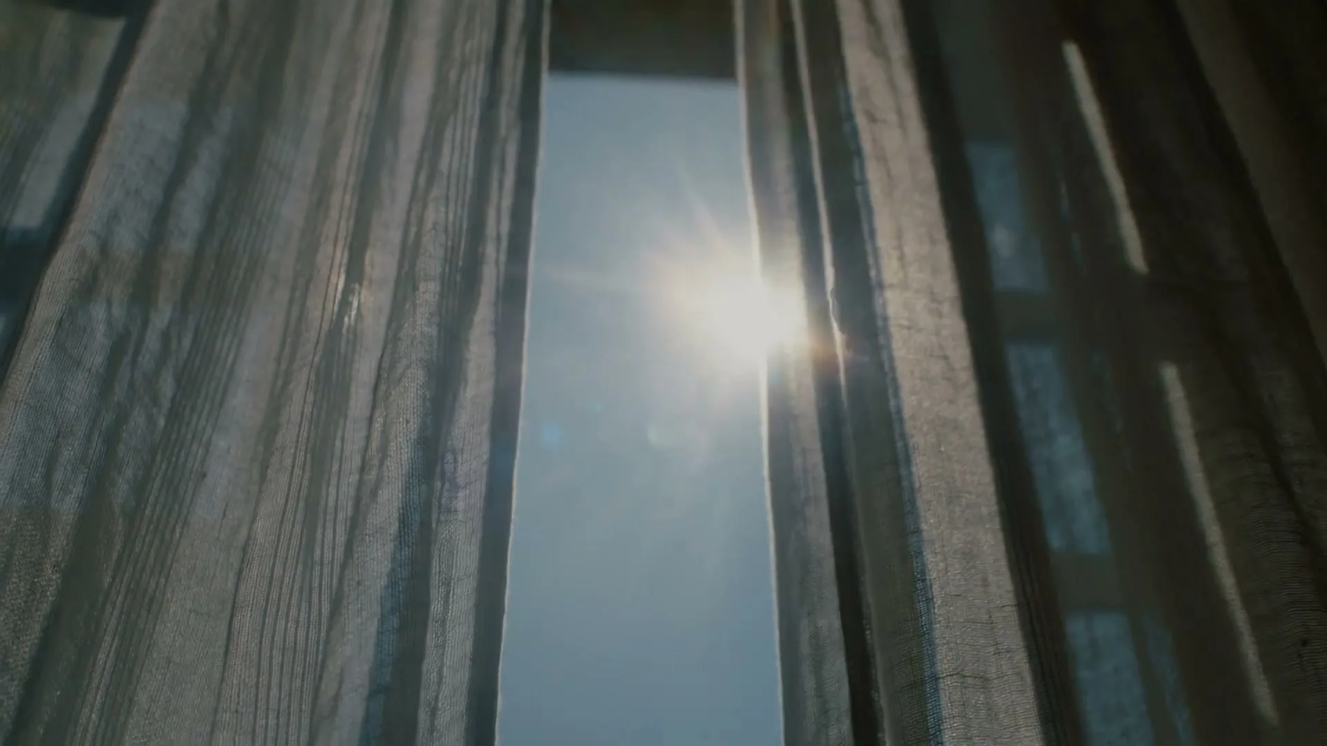 Sunlight streams through a gap in sheer curtains, creating a bright lens flare against a clear blue sky.