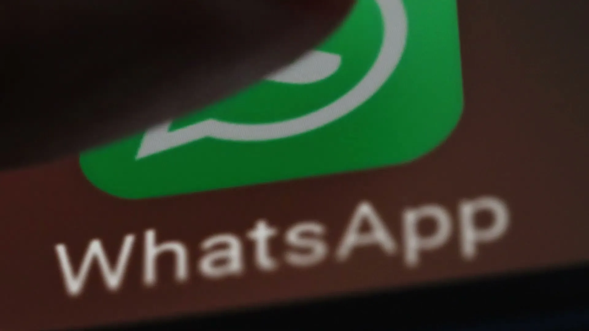 A finger taps the WhatsApp icon on a smartphone screen, showing the green logo and white text against a dark background.
